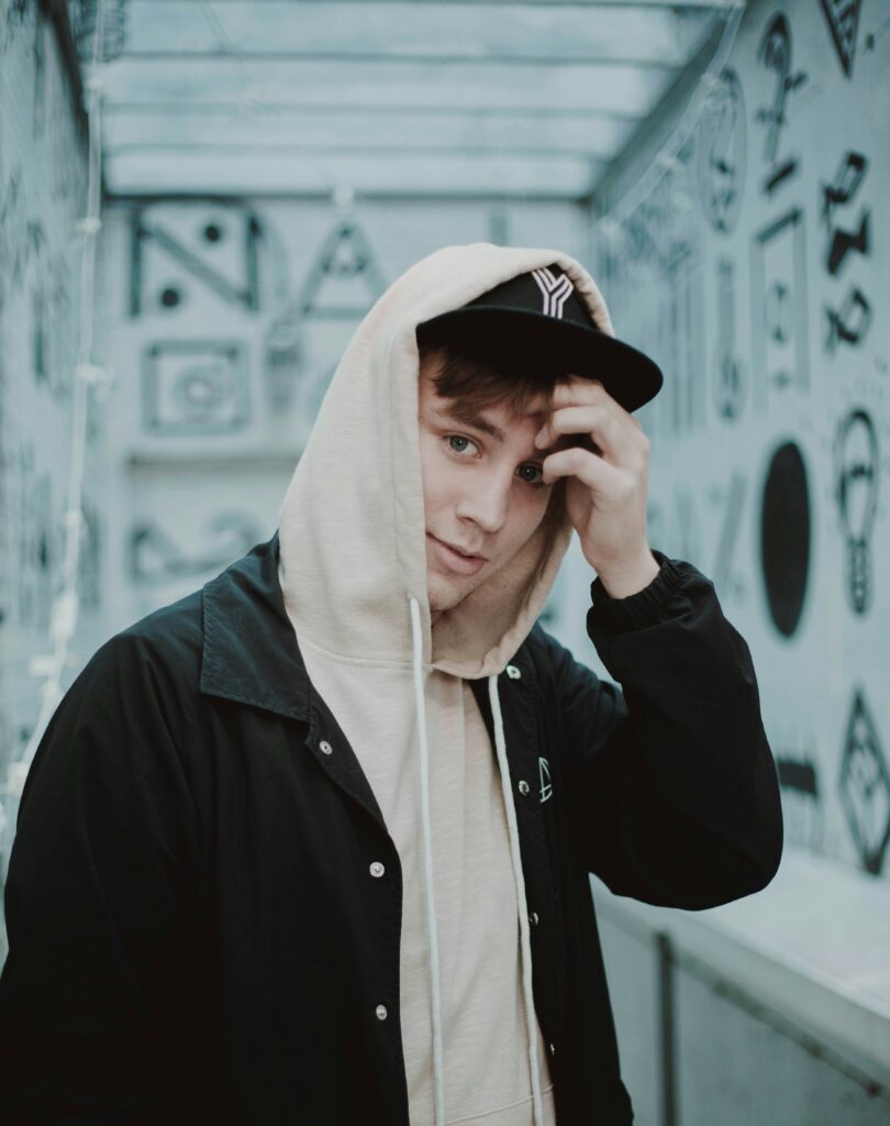 Nuori mies, jolla on lippis ja housu, katsoo kameraa käsi lähellä kasvojaan.